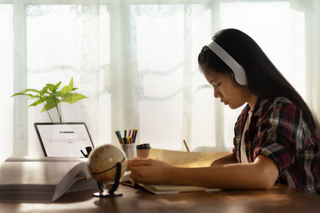 Female student wears headphones sitting at table to learning and note, using laptop for homework...