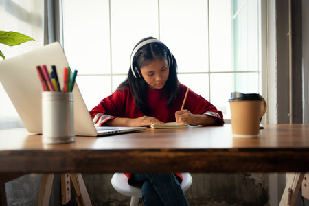 Female student wears headphones sitting at table to learning and note, using laptop for homework during covid19 pandemic and lock down, Social Distance, New life of Student on internet.の写真素材