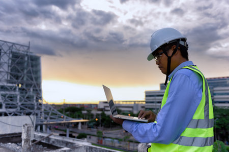 Civil Engineer wearing high visibility safety vest and helmet while using laptop looking to drawing detail on the terrace of decaying building at construction site. Surveyor for renovation concept.の写真素材