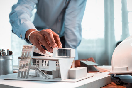 Hand of Asian architect-designer holding the part of the model of modern box house while thinking about concept of building and construction project in the future. Focusing on his hand holding model.の写真素材