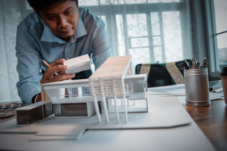 Asian architect-designer holding the part of the model of modern box house while thinking about concept of building and construction project in the future. Focusing on his hand holding model.の写真素材
