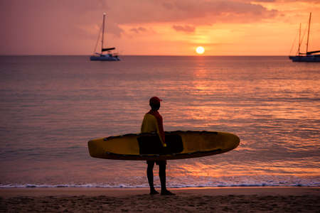 Silhouette of beach lifeguard standing alone with a surfboard on the empty beach while watching the...