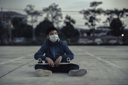 Tired Asian Kid Wearing Protective Face Masks Sitting Outdoors with Skateboard, Skateboarders on Street Keeping Social Distancingの写真素材