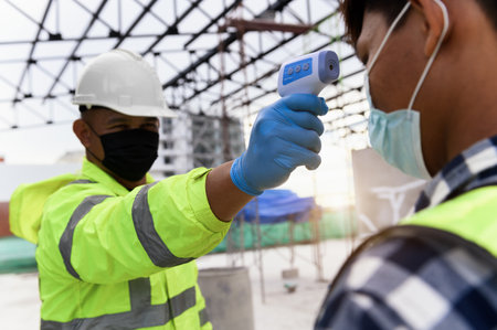 Construction foreman wearing protective face mask screening body temperature of worker with Infrared...