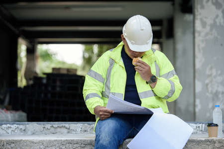 Civil Engineer sitting eats small bread in break time and reading building drawing with construction materials stock in background while hot weather at construction site during the day.の写真素材
