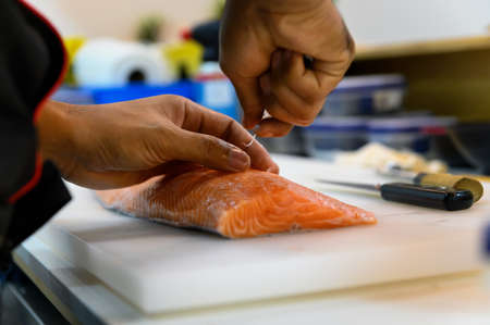 Fresh salmon fillet close-up on white cutting board table while fishbone removal with stainless steel tweezers âby chef in cooking room. Selection focus on salmon fillet with Soft focusing shootingの写真素材