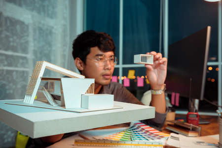 Architect-designer working at night with model of modern box house while thinking about concept of building and construction project before startup in the future.の写真素材