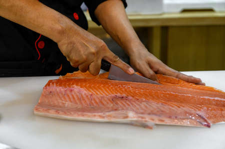 Fresh salmon fillet close-up on white cutting board table while filleting or slicing by chef in cooking room. Selection focusing on hand while slicing with Soft focusing shooting.の写真素材