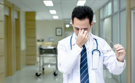 Asian senior doctor man wearing medical uniform standing while take off glasses with tired rubbing nose and eyes feeling fatigue and headache in hospital corridor. Stress and frustration concept.の写真素材