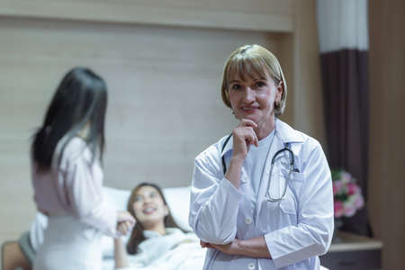 Caucasian senior woman doctor smiling while holding hand on chin and standing with background have the relatives visiting the patients who were treated improved.の写真素材