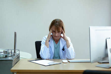 Old short blonde hair woman doctor with light blue shirt and white coat sitting on black chair in bright office in front of wireless computer stress and headache with many difficult case beside laptopの写真素材