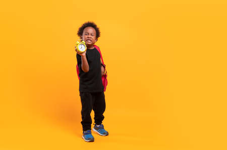 Black child boy 3 years, Students kid with school bag to hold alarm clock and showing time to enter the classroom with disgusted expression. Isolated portrait on yellow background with copy space.の写真素材