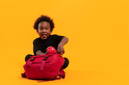 Black child boy 3 years, Students kid enjoy and stick out tongue as he was about to collect the apple, put them in the red school bag. Isolated portrait on yellow background with copy spaceの写真素材