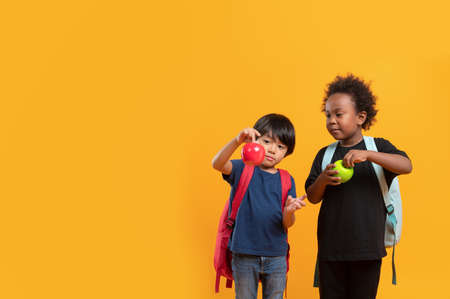 Two students 3 years, Asian and Black child boy of different nationalities carry a school bag and stand playing with apples isolated on yellow background with copy space, International student kid.の写真素材