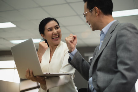 Office Asian female worker wear nice suit holding a laptop on left hand talking to colleague with smiling face raise hand to cheer up don't give up with colleague. Success business job and workingの写真素材