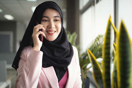 Female employee muslim wear pink suit with hijab stay in office behind her have many flower pot with morning light in background. She talking on phone with smiling face her phone on working day.の写真素材