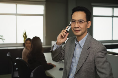 Smiling Asian businessman with glasses and suit holding smartphone observe employees in busy office, representative, postureの写真素材