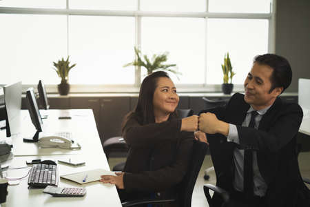 Asian Co-worker are bump their hands face to face with smiling face in office work area with many computers keyboards, mouses, house phone, Motivated to workの写真素材
