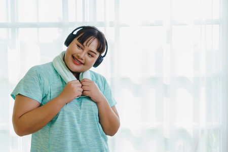 Portrait of Asian plus size female in sportswear with headphone doing body pose relax after exercising indoors to burn fat, making body strong, Healthy lifestyle at home.の写真素材