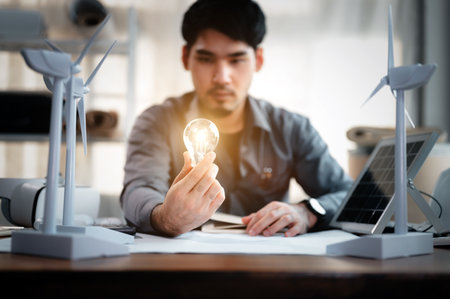 Energy engineer holding light bulb in modern office with solar cell sample and wind turbine model, Creative thinking innovative alternative energy design. Saving nature or ecology concept.の写真素材