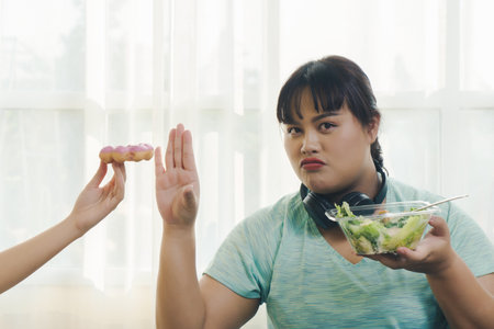 Fat woman Asian in sportswear using hand push out donut sweets. rejecting dessert or junk food with determined face while holding fresh salad in her hand. healthy lifestyle and diet concept.の写真素材