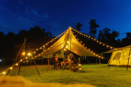 Outdoor camping tent with tarp or flysheet on grass courtyard and warm night light under dark blue...