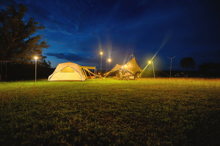Outdoor camping tent with tarp or flysheet on grass courtyard and warm night light under dark blue...