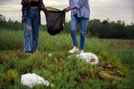 Closeup of plastic garbage at nature park with two female volunteers collecting garbage and plastic waste bag into garbage bags in background. Selective focus on plastic bags.の写真素材