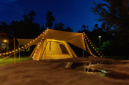 Outdoor camping tent with tarp or flysheet on grass courtyard and warm night light under dark blue sky twilight time, family vacation picnic on holiday relax, Overview of camping of family tourist.  **Focus**の写真素材