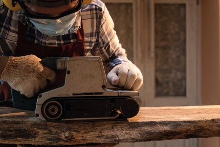 Carpenter use a hand-held electric sanding machine belt sander to level surface wood in carpentry workshop.の写真素材