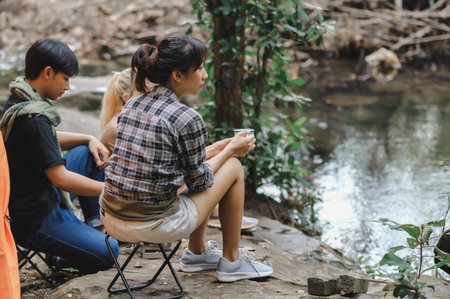 Young People Trekking Group Sitting Relaxed in the Rainforest Near a Natural Stream. Campsite Drinking Coffee and Water. Prepare Camping Meals for Groups on Trip.の写真素材