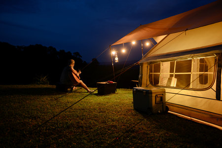 A person plays guitar beside a warmly lit tent at night, sitting under hanging lanterns on a grassy hill, creating a peaceful and cozy camping atmosphere with mountains in the background.の写真素材