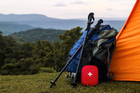 Close up a Backpack, trekking poles, and a red first aid kit rest beside an orange tent on a grassy hill, Rest and Recovery heal body with mountains a cloudy sky in the background, outdoor adventureの写真素材