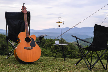 An acoustic guitar, camping chairs, a lantern, and a mug are set up on a grassy hill with a mountainous backdrop, creating a peaceful outdoor retreat.の写真素材