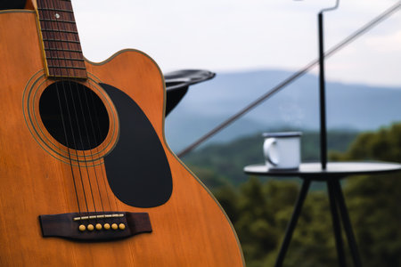 Close up of a wooden acoustic guitar is positioned outdoors next to a small table with a mug, against a backdrop of mountains and greenery, creating a peaceful and relaxing atmosphere.の写真素材