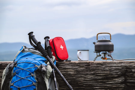 A camping setup with a backpack, trekking poles, a first aid kit, a mug, and a teapot resting on a log, overlooking a serene mountain view. The scene highlights the essentials for an outdoor adventure.の写真素材