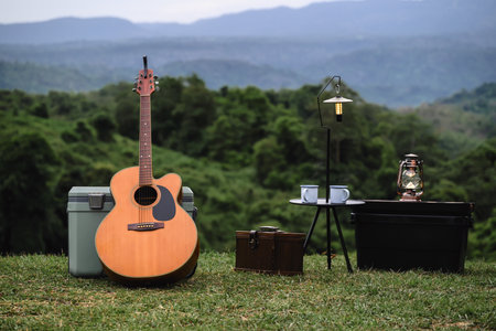 An acoustic guitar stands beside a cooler and camping gear on a grassy hilltop, with a lantern and coffee mug placed on a small table. The lush mountains in the background.の写真素材