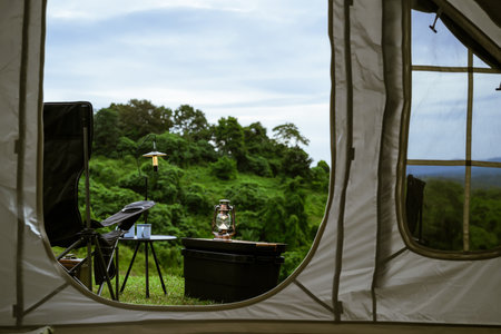 A peaceful view from inside a tent shows camping gear, including a lantern and a chair, set on a grassy hill with lush greenery in the background, offering a serene outdoor experience.の写真素材