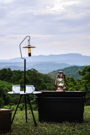 A small camping setup with two mugs, a lantern, and a table overlooks a beautiful mountain landscape. The peaceful scene, surrounded by lush greenery, offers a serene outdoor retreat.の写真素材