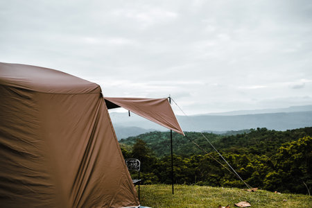 A camping tent is pitched on a grassy hill, overlooking a lush green forest and distant mountains under a cloudy sky, creating a serene and scenic camping atmosphere.の写真素材