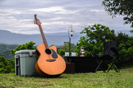 An acoustic guitar stands beside a cooler and camping gear on a grassy hilltop, with a lantern and coffee mug placed on a small table. The lush mountains in the background.の写真素材