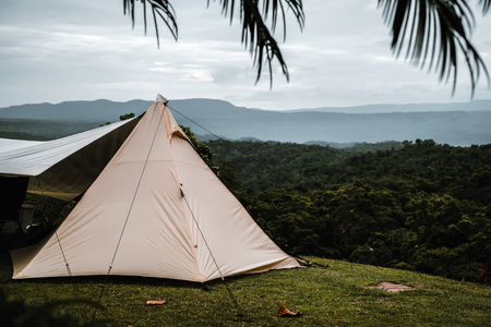 A triangle-shaped tent is pitched on a grassy hill, overlooking a lush green forest and distant mountains under a cloudy sky, creating a serene and scenic camping atmosphere.の写真素材