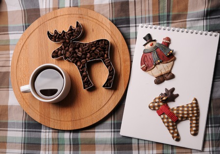Cup of coffee, coffee beans in the shape of elk on wooden board, notebook with figures of deer and...
