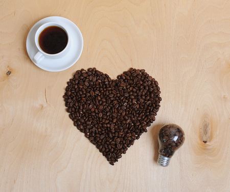 Large heart made of coffee beans, cup of coffee and bulb with coffee beans inside on a light wooden background. Top viewの写真素材