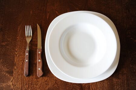 Empty white plate with fork and knife on a dark wooden brown tableの写真素材