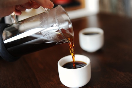 Hand pours coffee from a glass jug into a white espresso cup without a handleの写真素材