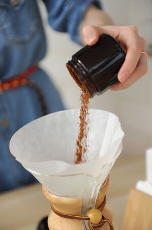 Pouring freshly ground coffee from glass grinder container into paper filter to brewing close up