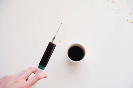 A typed espresso in a medical syringe in hand over a cup. Caffeine addiction symbol. White...