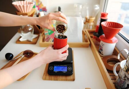 Alternative brewing. Pouring coffee beans into red manual coffee grinder on scale. Third wave specialty aestheticsの写真素材