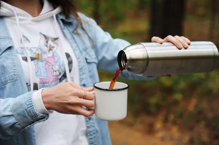 Coffee in travel. Girl pouring hot black coffee from tumbler in white metal cup outdoor. Close upの写真素材